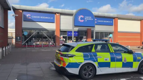 BBC A police car outside the Cancer Research UK store. A cordon has been set up around the building.