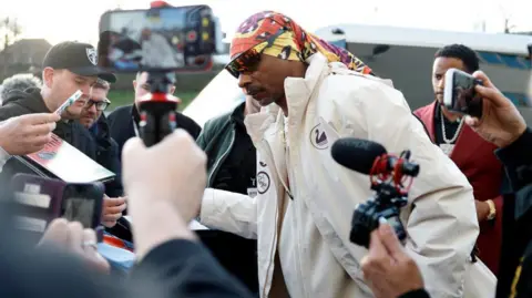 Reuters A man wearing sunglasses and a Swansea City Snoop Dog white coat is surrounded by members of the media holding phones and cameras as he arrives at the ground.