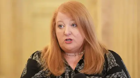 Naomi Long photographed mid-speech as she talks to reporters.  She is a woman with shoulder-length straight ginger hair.  She is wearing a black and white patterned blouse and a necklace with a silver clasp.
