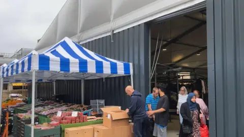 Elizabeth Baines / BBC A metal shipping container houses market traders, outside stalls are set up with blue and white striped canopies