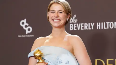 Jessie Buckley, a woman with short, blonde hair, poses with her Golden Globe award. She is wearing an off the shoulder silver/pale blue gown. 