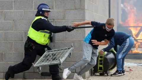 Reuters  A police officer in riot gear is holding a baton out towards a young man in a t-shirt and trousers, who is wearing a face mask and moving away from the officer. In the background a man in tracksuit bottoms and a blue top can be seen lifting a black container. Behind him there are large flames.