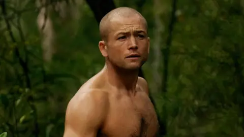 Egerton topless in a forest. He also ahs a shaved head
