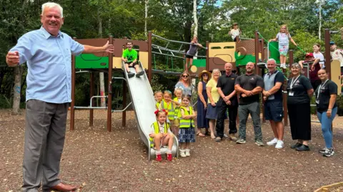 BBC/Marc Gaier A man with short grey hair and wearing a short-sleeved blue shirt and light brown trousers is smiling with his hands raised at either side of him as she stands next to a group of adults and children who are next to play equipment. The children and adults are all smiling.