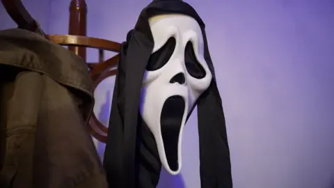 A mask from the horror film Scream hangs off a hat stand in the corner of a room with purple shadows on the walls