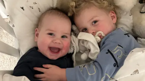 Smiling baby in a dark top lying down on a cushion being hugged by his older brother who has a dummy in his mouth 