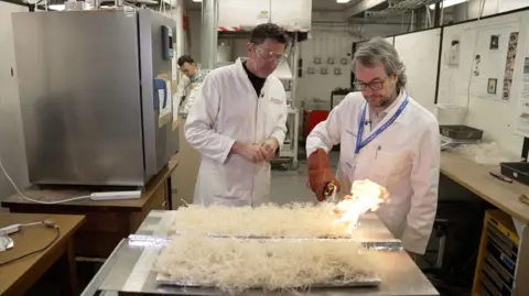BBC / Kevin Church A professor with grey hair with a pair of glasses wearing a white overall coat is igniting dry straw on a steel plate with a fire burner in a laboratory. Justin Rowlatt - also wearing a white overall coat and a pair of safety glasses - is looking on next to the professor.