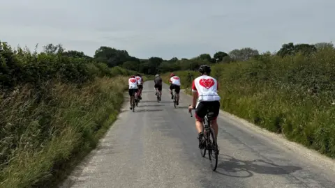 Wolverhampton Coronary Aftercare Support Group A group of people on bikes with their backs to the camera. They are cycling on a road, with bushes on both sides of the road.