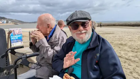 Supplied Image of John Crouchley, wearing a flat cap and a blue fleece and sat by the beach.