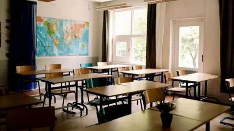 Photo of empty classroom with map of the world on the wall at the back. The classroom is tidy and all the chairs are tucked into the tables. There are large windows with trees showing on the far side. 