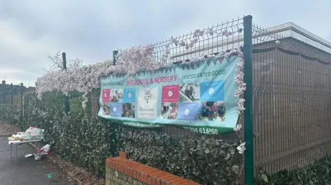 Alice Cunningham/BBC Another view of the side of Blossom Preschool and the banner that sits on its fence. A table sits outside the site with free food on offer.