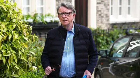 Peter Mandleson outside walking. He is looking at the camera and wearing a blue shirt and a navy coat