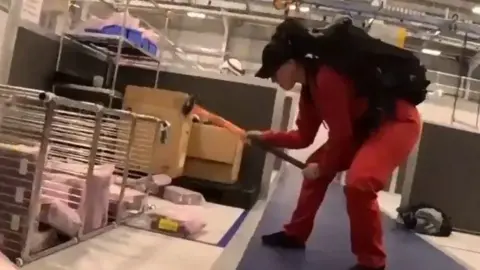 Palestine Action A person wearing a red jumpsuit and black cap, using a sledgehammer to damage equipment inside the factory.
