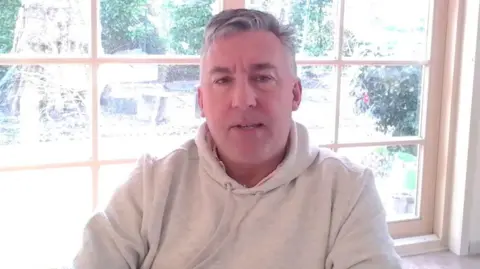 A man in a light coloured sweatshirt and a window behind.