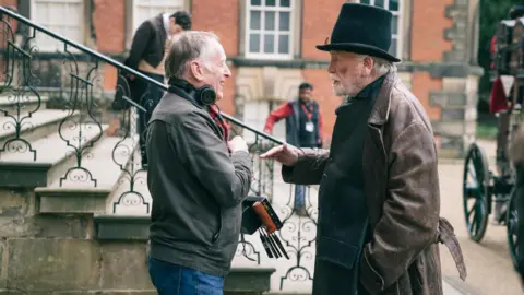 An outdoor scene set in front of a large, historic red‑brick building with tall white‑framed windows and decorative stone detailing around the entrances. In the foreground, Stone and Cosmo are standing face to face in conversation near the bottom of a short flight of stone steps with ornate black iron railings. Cosmo is wearing a dark coat and a tall black hat, while Stone is dressed more casually and has a pair of headphones resting around his neck while holding a small piece of equipment.