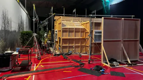 Josh Parry / BBC An exterior of a film set, propped up by scaffolding. It's a wooden box with windows and outside the windows are fake trees and shrubs. The floor is covered in cables and filming equipment.