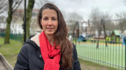 Emily Jennings. She is wearing a red scarf and a dark jacket. She has long brown hair. She is standing in front of a blurred background of a playground. 