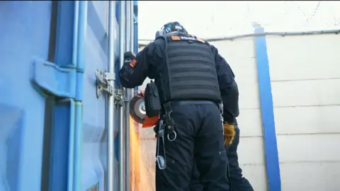 Law enforcement officers with black vests and heavy duty tools are cutting into a large blue storage unit container. The machinery is sparking as they carve through the metal.