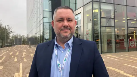 Kate Bradbrook/BBC Jason Smithers wearing a blue shirt and blue jacket standing outside The Cube in Corby