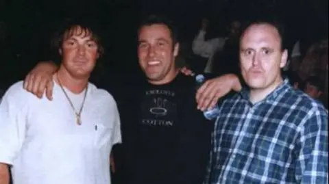 Bernard O'Mahoney Tony Tucker, Pat Tate and Bernard O'Mahoney with their arms around each other in a club. Tucker is wearing a white T-shirt and a gold chain. Tate is wearing a black sweater and holding a water bottle. Mr O'Mahoney is wearing a blue and white checked shirt.
