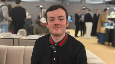 Carl Player está sentado en un sofá durante la venta. Lleva una blusa negra con cuello rojo y gris y luce orgulloso.