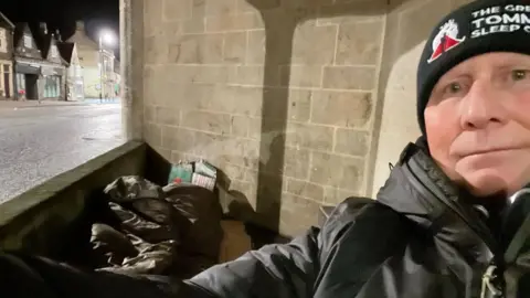 A man in a black coat, wearing a black wool hat, sits in what appears to be a shelter. It's dark. There is a sleeping bag behind him.