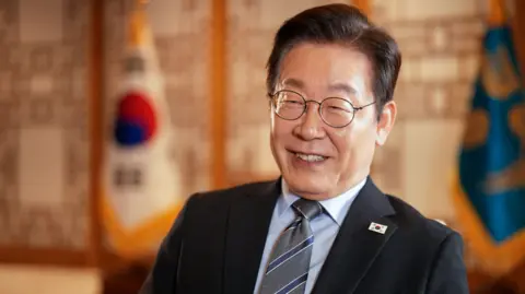 BBC/ Hosu Lee President Lee Jae Myung sits in a chair in a black suit in his office in Seoul. Behind him are two flags. 