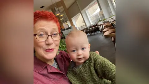 Cancer Research UK Self taken portrait photo of Fiona Carey with her young grandson. She has blue eyes and is looking at the camera. Her grandson, who also has blue eyes, is sat on her knee wearing a khaki-green jumper. They appear to be sat in a canteen or restaurant, as there are tables and chairs in the background. 