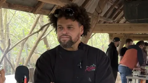A man with curly hair, sunglasses and a dark top in an outdoor area with a roof over the top