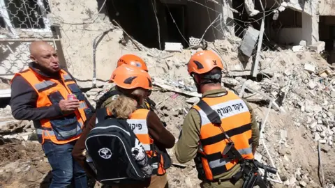Emergency workers wearing orange hi vis and helmets gather in front of strike damage at the site in Tel Aviv on Tuesday