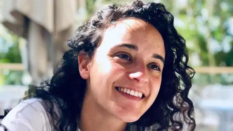 PA Media Emily Damari, a young woman with curly dark brown hair wearing a white t-shirt, smiles.