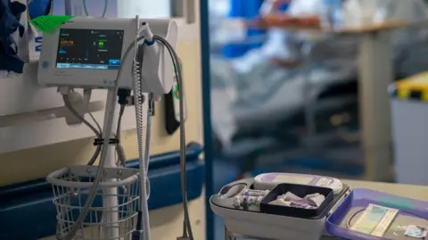 PA Media Hospital equipment with a monitoring screen and a bed with what appears to be needles and further monitoring equipment on it. A hospital room, including a bed, can be seen in the blurred background. 