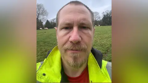 Chris Haffenden, seen in a selfie, stands outside with a field, sheep and trees behind him. He has a beard and is wearing a hi-vis jacket with a red top.