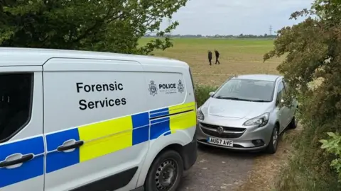 Steve Hubbard/BBC A police van and a car at the entrance to a field. The van says 'forensic services' on it. In the background, there are two people in dark clothing walking across the field.
