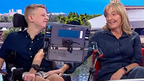 A young man in a wheelchair and with a tablet in front of him is next to a woman in a denim dress on a red sofa in a studio. She is holding on to his knee.