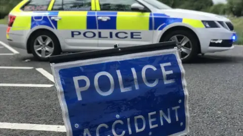 A blue 'Police Accident' sign in close-up on a road, while a stationary police car sits behind it