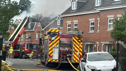 Two fire engines and firefighters attending to a house fire. There is smoke pillowing above the roof. There is also a white car parked by a gate. 