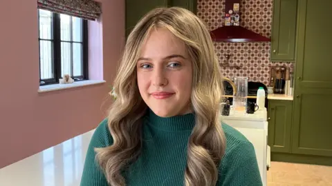 A young woman with long blonde hair, wearing a teal jumper. She is staring into the camera lens and is stood in a kitchen, with pink walls and green cupboard doors, a white island and patterned tiling 