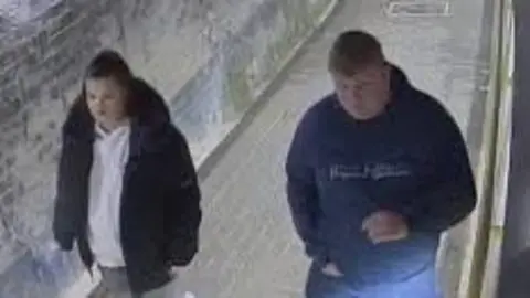 Gloucestershire Constabulary CCTV image of two teenagers walking side by side. The girl on the left is wearing grey leggings and a black fur-hooded coat and the boy on the right in a navy McKenzie tracksuit with blond hair.