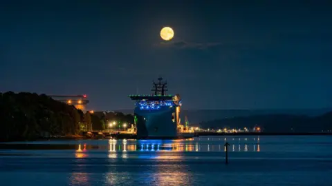 Tim Berrall A large vessel moored on calm water at night beneath a bright full moon, with colourful dock lights reflecting across the water’s surface.