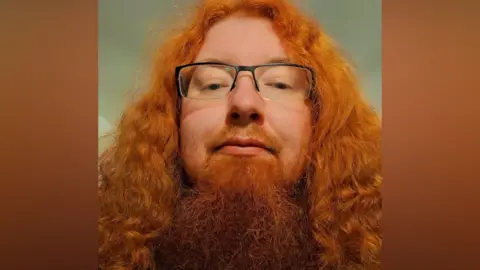 Adam Sharples Adam Sharples has long red hair and a long red beard. He is wearing glasses. 