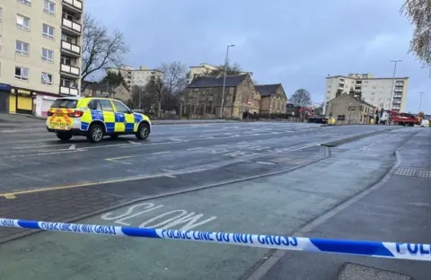 BBC/Adam Laver A police vehicle parked in the middle of a deserted street with blue and white police tape in the foreground.
