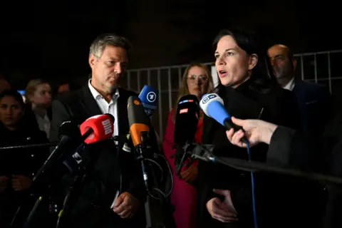 Reuters Economy Minister Robert Habeck wearing a suit and shirt and Foreign Minister Annalena Baerbock wearing a black coat stand in front of microphones to speak to the media.