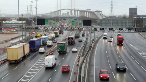 Cars, lorries and vehicles queuing to use the Dartford Crossing between Kent and Essex. There are several lanes of traffic. The large QEII bridge is pictured in the background.