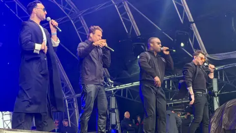 Boy band Blue on stage. All four members have a microphone. They are backlit by blue lighting