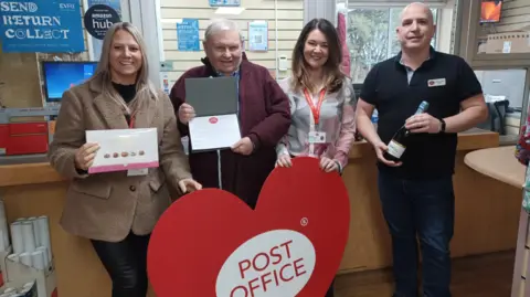 Left to right: Post Office area manager Nicola Ballard, Roger Honey, Post Office regional manager Debbie Mickleborough and Jamie Davey, who has worked at the branch for 26 years.
The group are standing inside the Post Office branch. Mr Roger is holding the award and is standing in front of a large red and white cut out a heart-shape which reads Post Office in the middle.

 