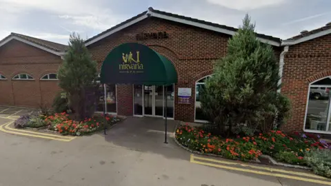 A Google Maps picture of the spa's entrance, which is in a one-storey building with large conifers on either side of a green covered area with Nirvana Spa written on the the front it.