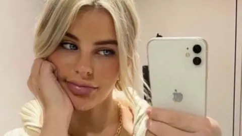 The photo shows Paris Wilson taking a mirror selfie indoors. They are holding a white iPhone with a dual‑camera setup in their right hand, and they are wearing a chunky striped knit top with a zip collar. Their hair appears to be light blonde and tied back loosely. A gold chain necklace is visible around their neck, and they are wearing a gold ring on their right hand. The setting looks like a simple, neutral-toned room, possibly a bedroom or changing area.