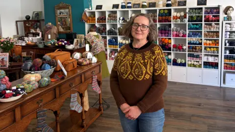 LDRS Venetia Strangways-Booth is wearing a brown jumper decorated with yellow fish on its upper half, as well as jeans. Behind her a wide range of different wools are on shelves.