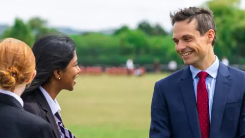 Joel Osborn Photography/Pate's Grammar School A photo of headteacher Dr Richardson smiling while talking to two pupils. He has short, dark hair, and is wearing a red tie, light blue shirt and blue suit.
